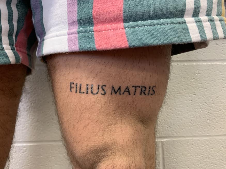 Luke Mathwigs tattoo says, Filius Matris which is a Latin phrase which translates to ‘Mother’s son’.