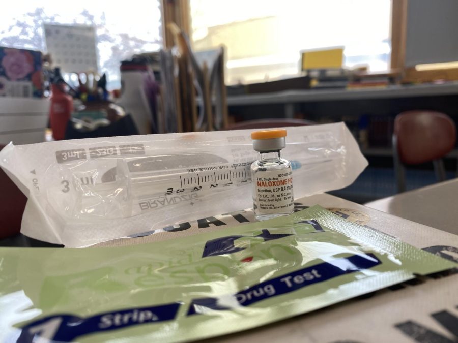 Pictured are a fentanyl test strip, a vial of naloxone, and a syringe, which are all measures that can be taken to prevent a drug overdose.